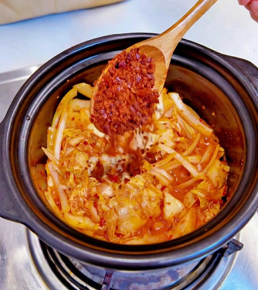 Un cucchiaio di legno aggiunge peperoncino rosso in fiocchi in una pentola nera contenente kimchi e cipolle.