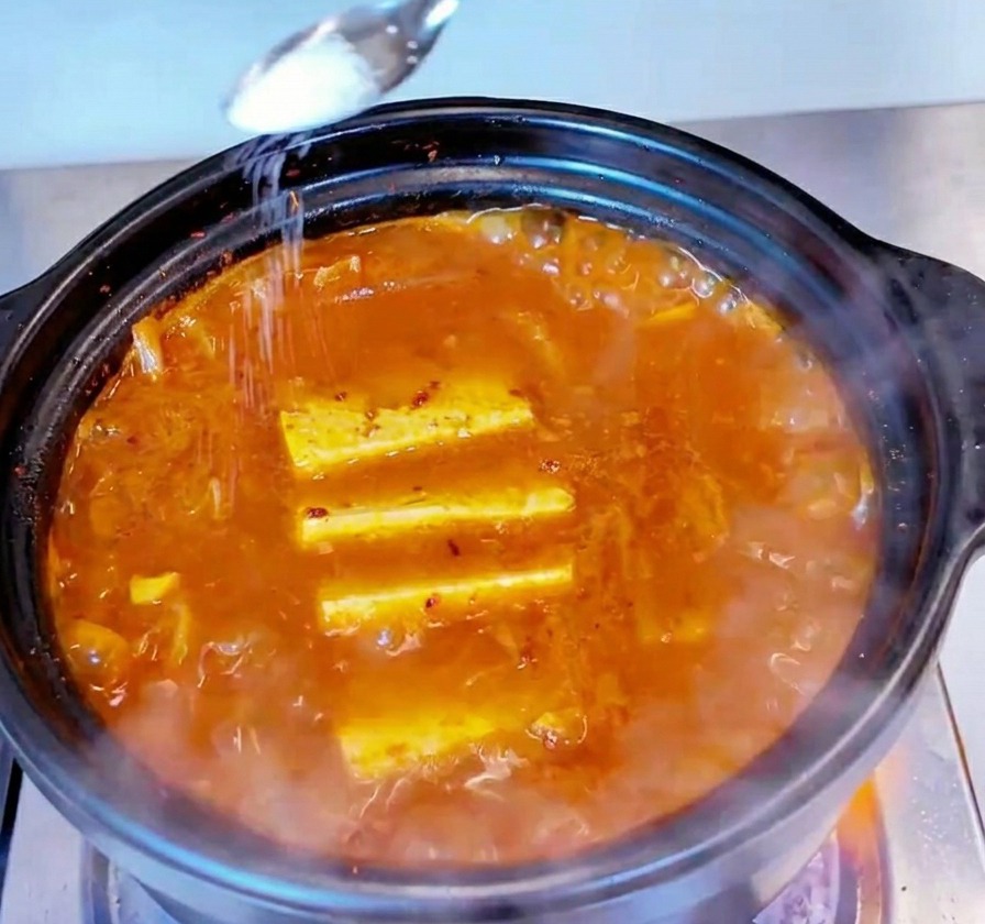 Un piccolo cucchiaio di metallo sparge del sale bianco sulla superficie di uno stufato di kimchi e tonno bollente.