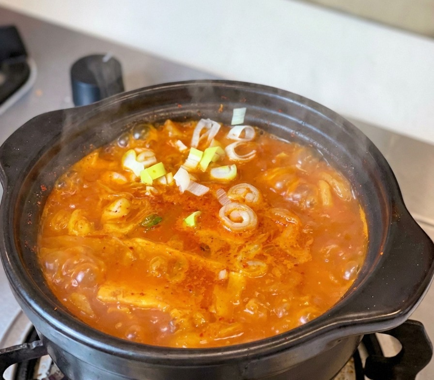 Uno stufato di kimchi e tonno bollente in una tradizionale pentola di terracotta nera sul fuoco, guarnito con cipollotti freschi e fette di peperoncino.