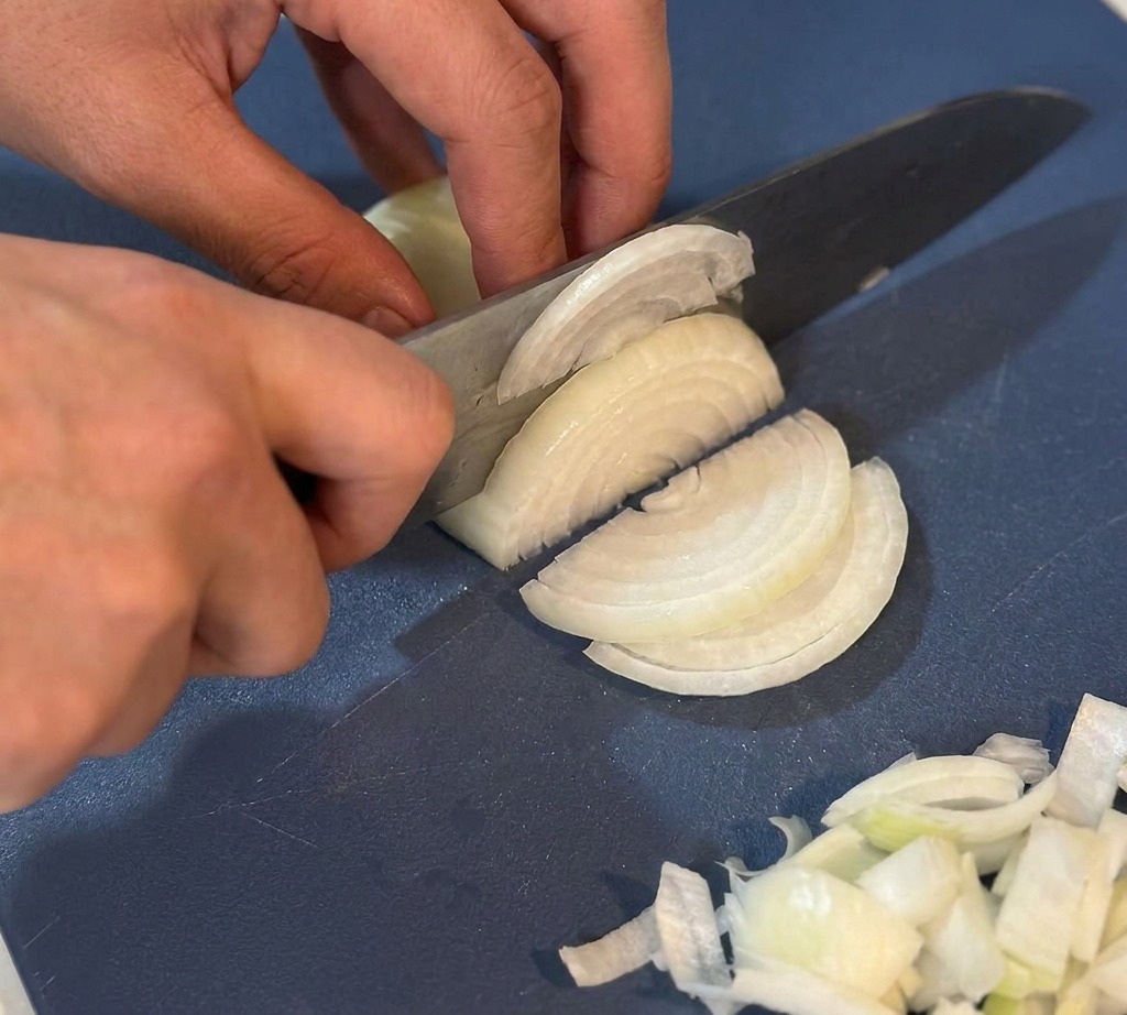 Mani che usano un coltello da chef per affettare sottilmente una cipolla bianca su un tagliere blu.