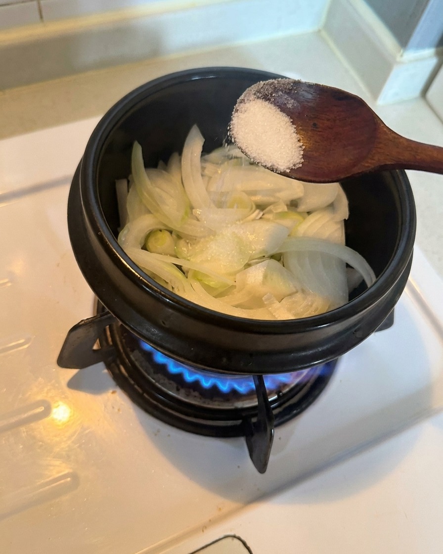 Cipolle bianche affettate in una pentola condite con un cucchiaio di zucchero bianco su un bruciatore a gas acceso.