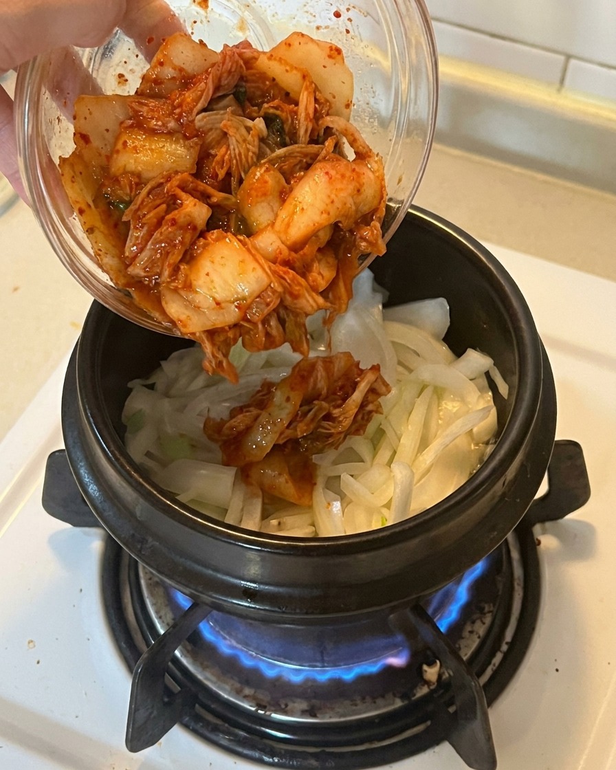 Inserimento di una ciotola di kimchi tritato di un rosso vivace in una pentola con cipolle e cipollotti soffritti.