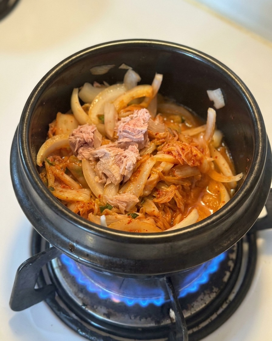 Pezzi di tonno in scatola adagiati su un letto di kimchi e cipolle soffritti in una pentola nera.