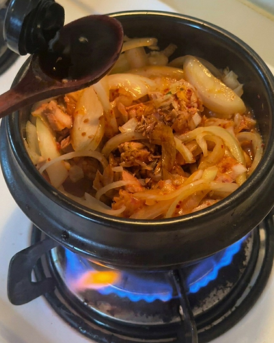 Un cucchiaio di legno versa della salsa di soia scura in una pentola di terracotta nera contenente cipolle, tonno e kimchi saltati in padella.