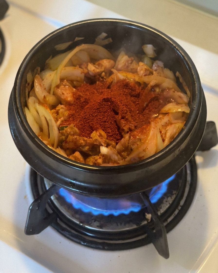 Una generosa manciata di gochugaru rosso brillante sopra il kimchi e le cipolle saltati in una pentola di pietra nera su una fiamma a gas.