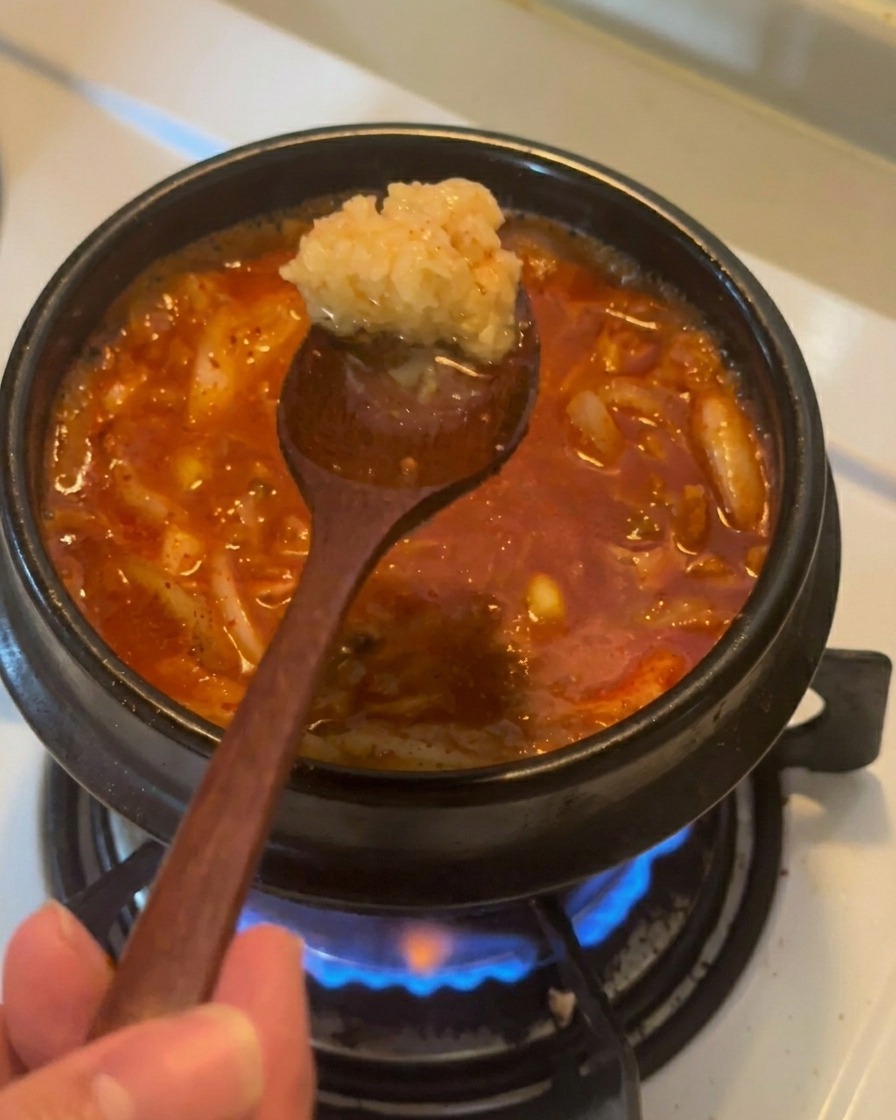 Un cucchiaio di legno tiene una porzione di aglio tritato sopra uno stufato di kimchi e tonno rosso che bolle.