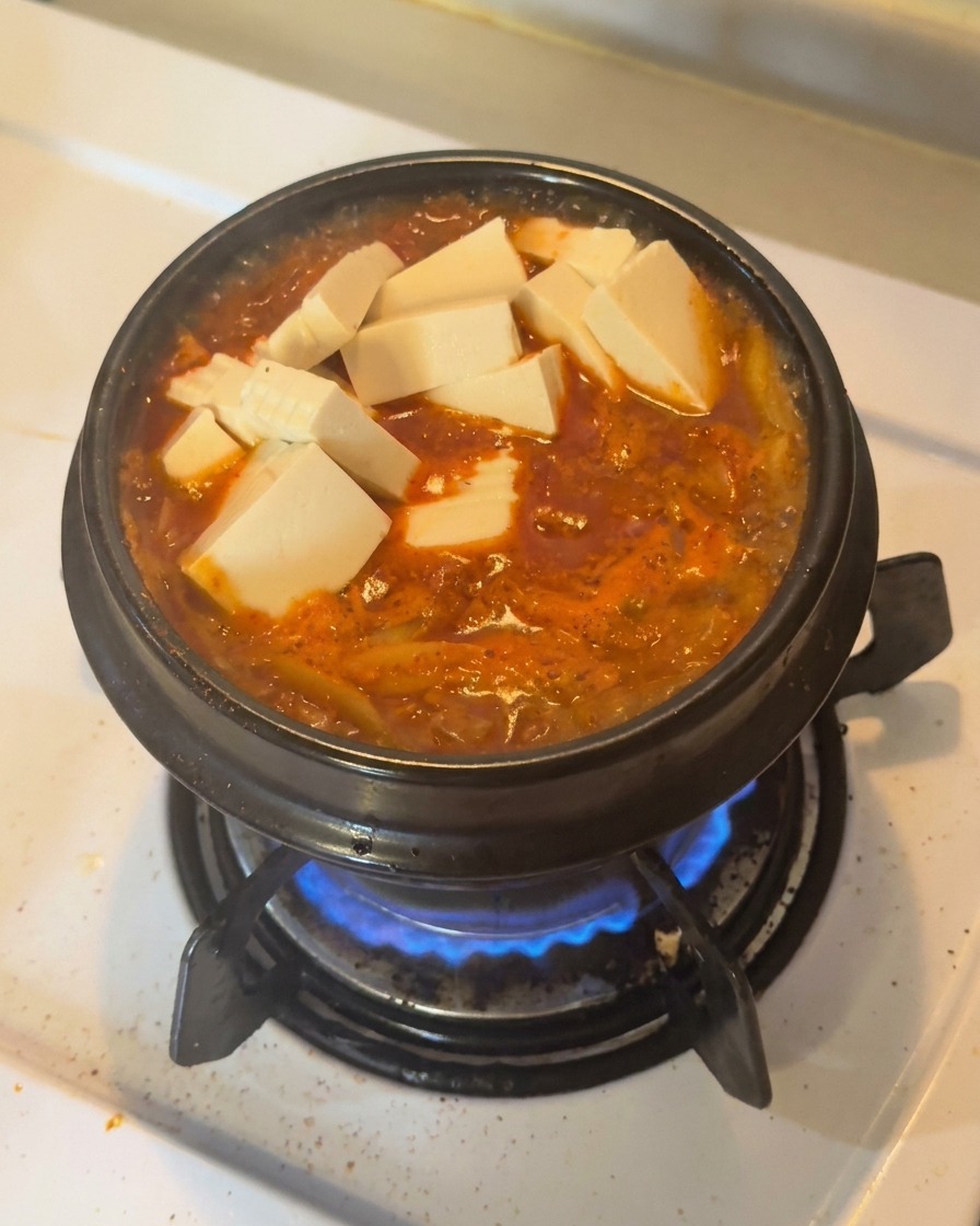 Cubetti di tofu bianco fresco e fette di peperoncino verde disposti sopra uno stufato di kimchi rosso che bolle in una pentola di terracotta nera.