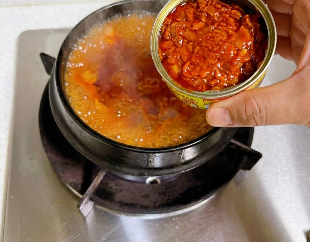 Una mano che tiene una scatoletta di tonno aperta e ne versa i pezzi nel brodo rosso di kimchi che bolle in una pentola di pietra.
