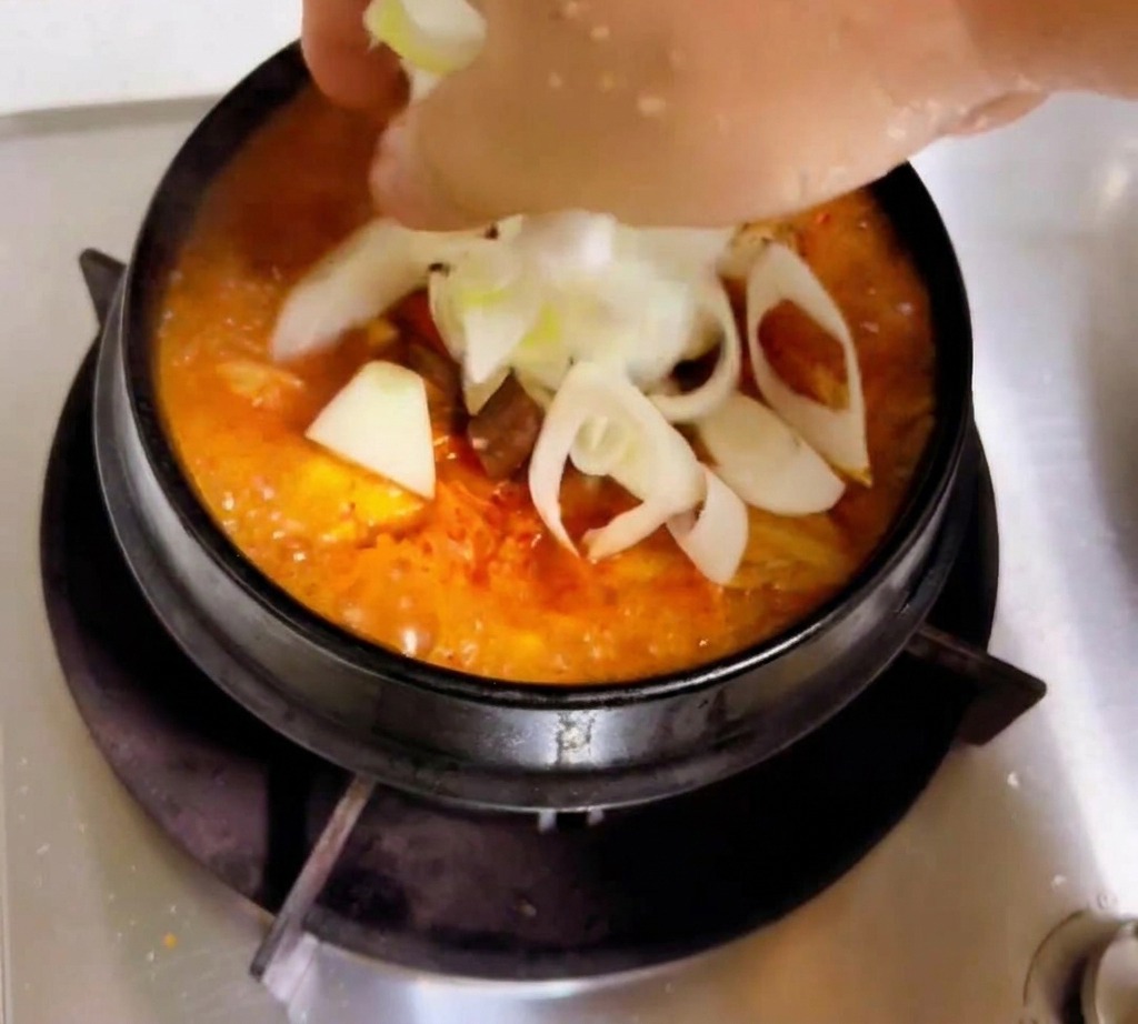 Una mano che aggiunge cipolle bianche affettate e cipollotti a uno stufato di kimchi arancione-rosso che sobbolle.