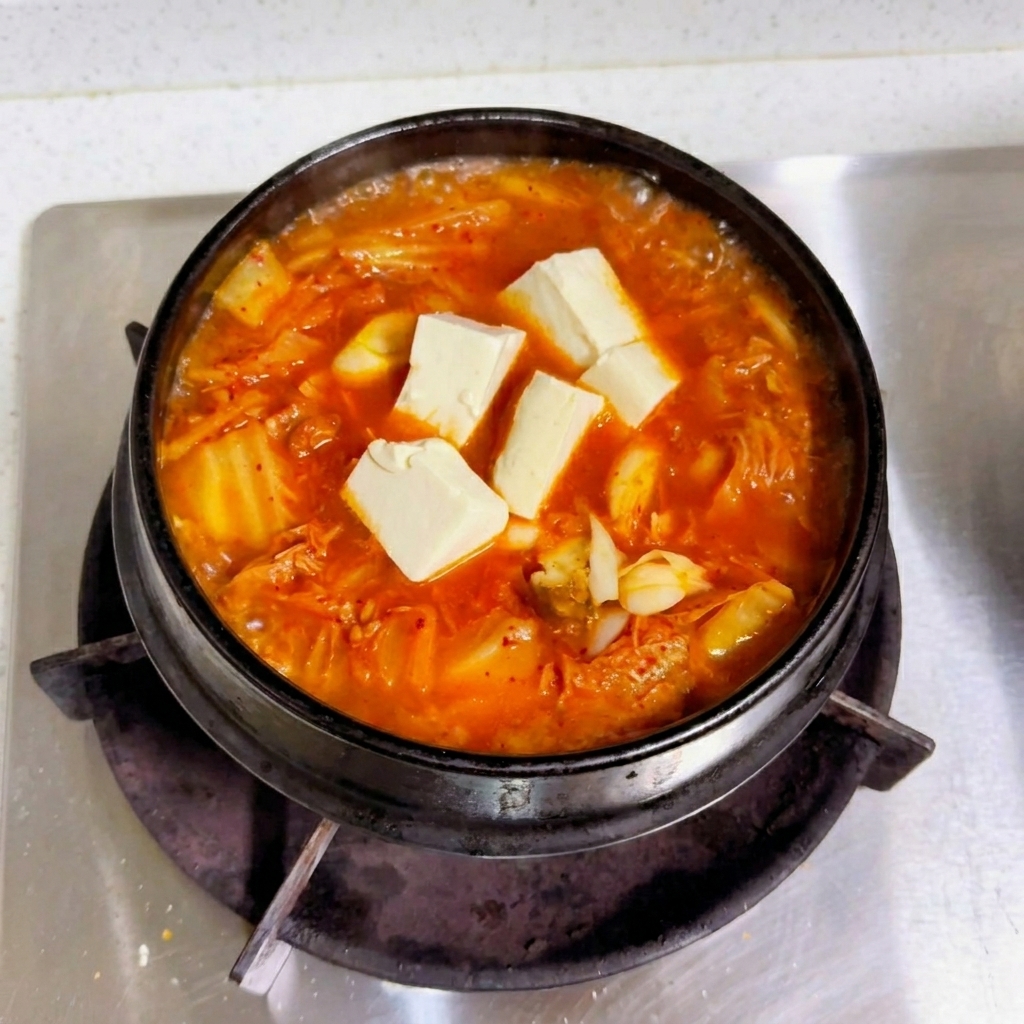 Cubetti di tofu bianco fresco adagiati sopra un brodo rosso di kimchi che bolle in una pentola nera.