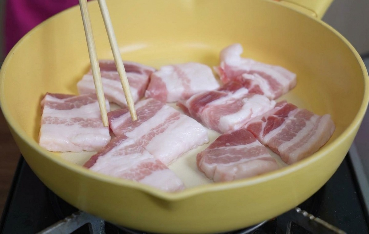 Fette spesse di pancetta di maiale cruda disposte in una padella di ceramica gialla, sistemate con le bacchette.