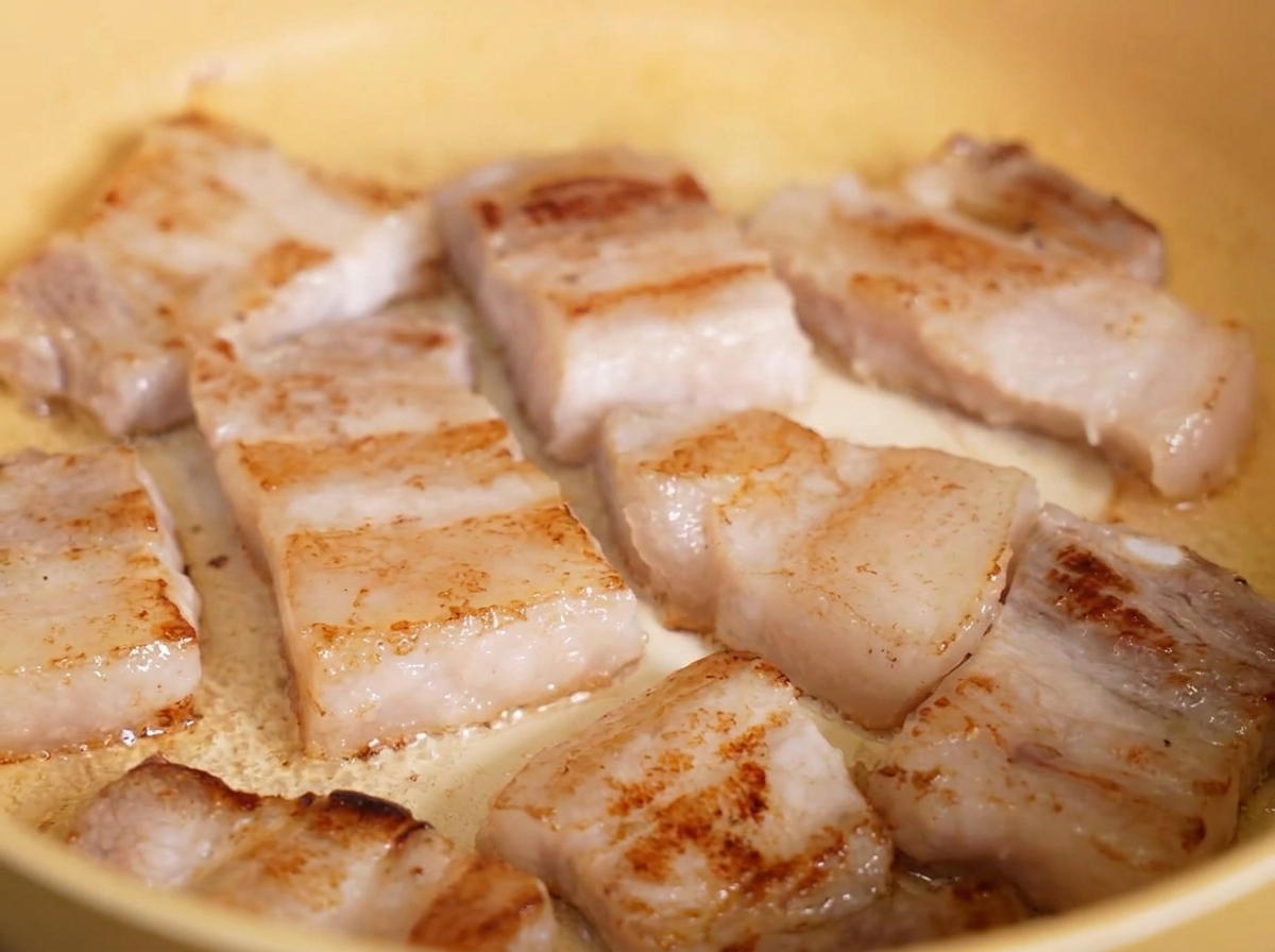 Fette di pancetta di maiale fritte, croccanti e dorate che sfrigolano nel grasso fuso allinterno di una padella gialla.