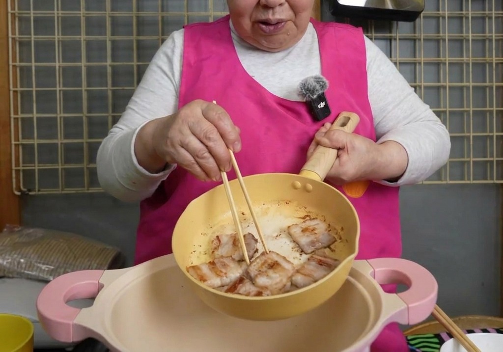 Pezzi di pancetta di maiale dorati che vengono spostati con le bacchette da una padella gialla a una pentola rosa più grande.