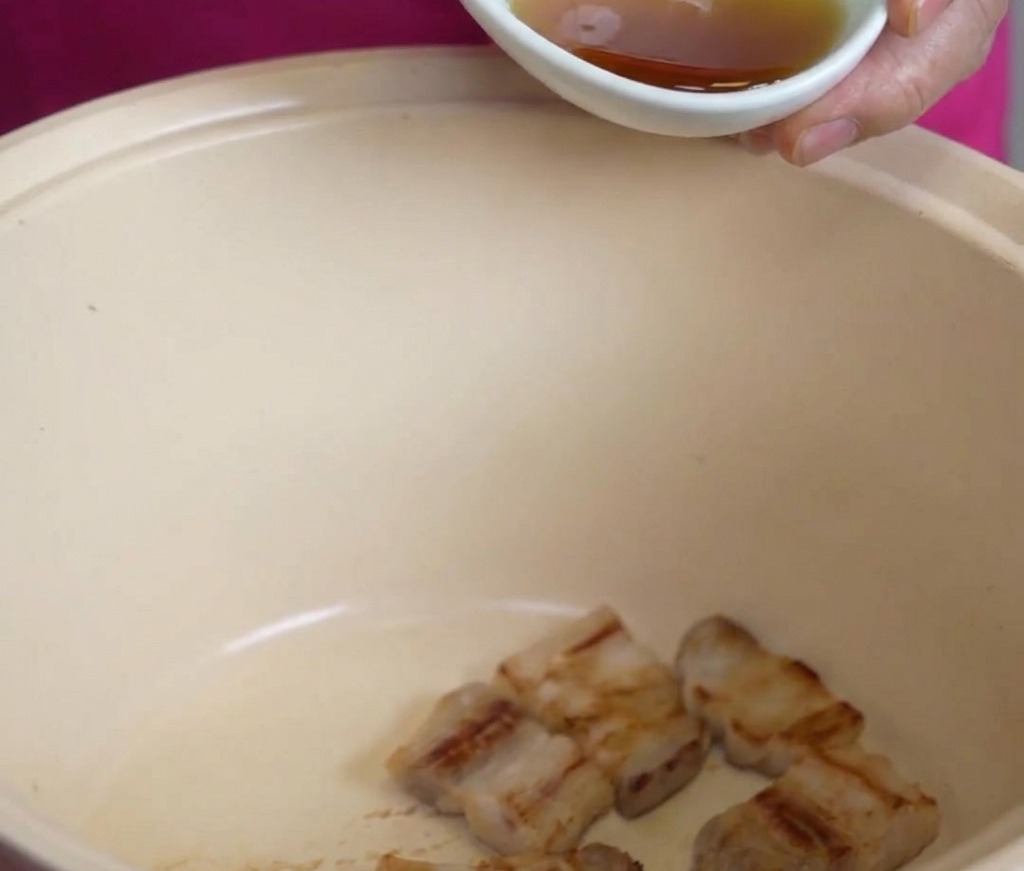 Una piccola ciotola bianca che versa olio di sesamo dorato sui pezzi di pancetta di maiale fritti in una grande pentola color crema.