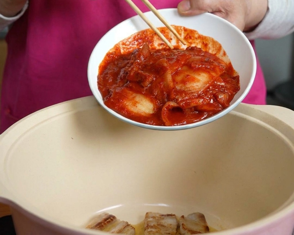 Una mano che usa le bacchette per far scivolare una ciotola di kimchi rosso piccante in una pentola contenente la pancetta di maiale.