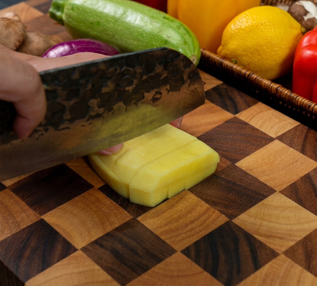 Un coltello da cucina che taglia una patata gialla pelata per creare cubetti uniformi su un tagliere di legno.