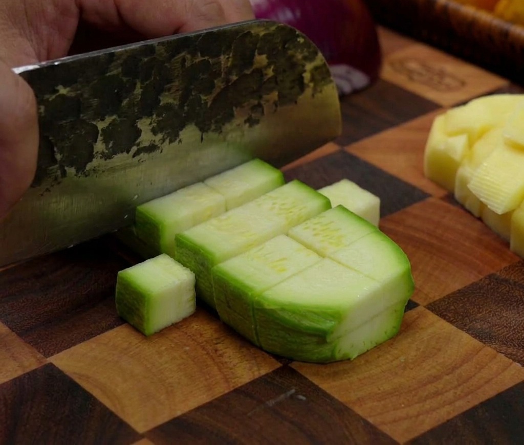 Una mannaia che taglia una zucchina verde, dividendola in piccoli pezzi rettangolari su un tagliere di legno a scacchi.