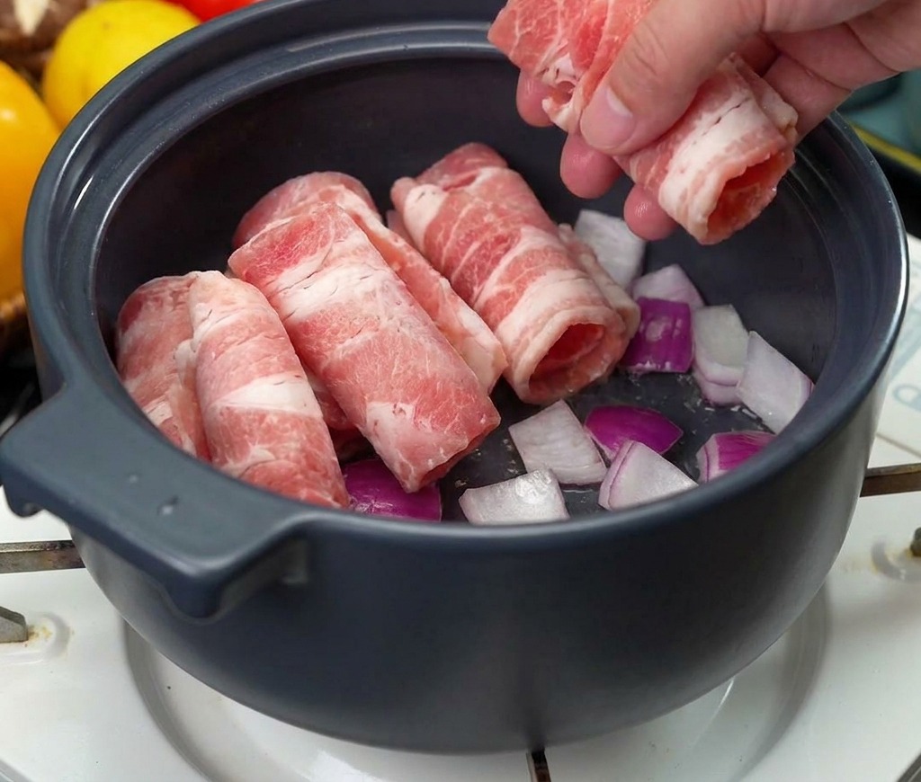Una mano che adagia diversi rotolini di carne di manzo cruda tagliata sottile in una pentola sopra un letto di cipolle soffritte.