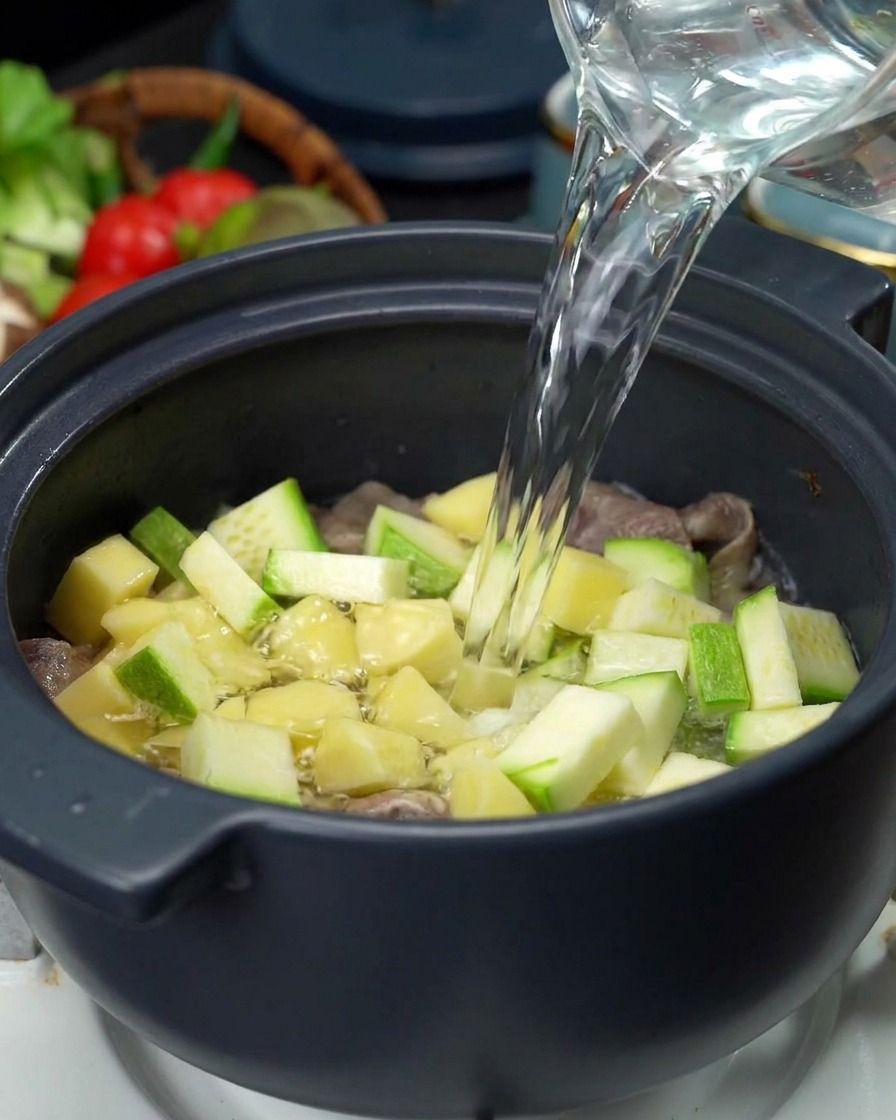 Un flusso di acqua limpida versato da una caraffa di vetro in una pentola colma di carne, patate e zucchine.