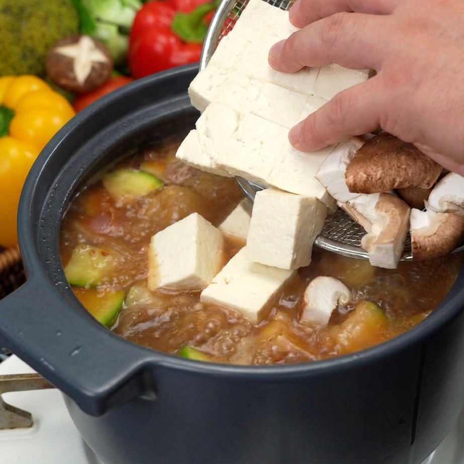 Una mano che usa una schiumarola per aggiungere cubetti di tofu bianco fresco e fette di funghi shiitake marroni in una pentola di zuppa bollente.