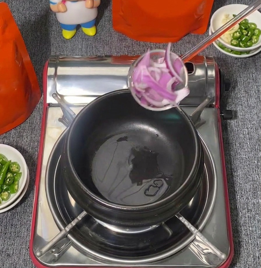 Un mestolo pieno di cipolle rosse affettate viene calato in una pentola di ceramica nera su un fornello portatile.