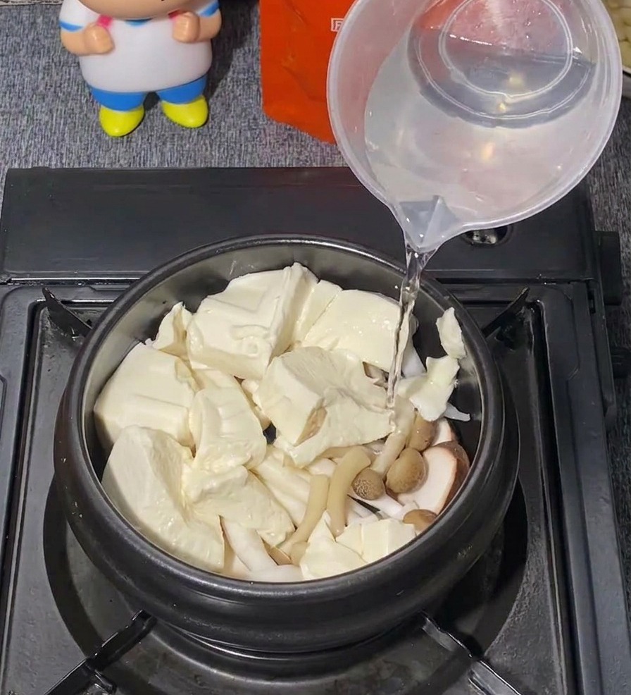 Acqua limpida versata da un misurino di plastica in una pentola nera piena di tofu e funghi.