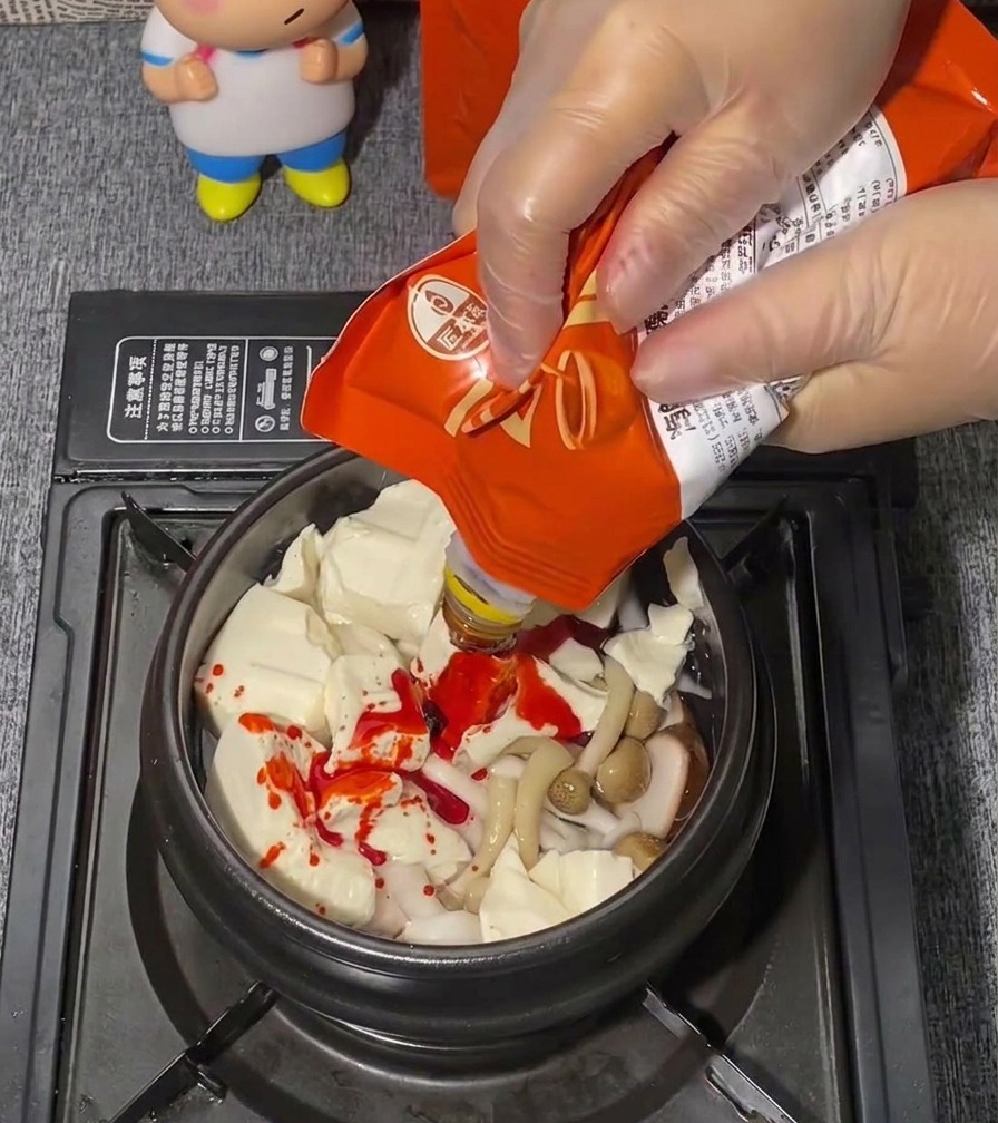 Una mano con un guanto di plastica trasparente spreme una pasta di condimento rosso brillante da una bustina su un letto di tofu tenero.