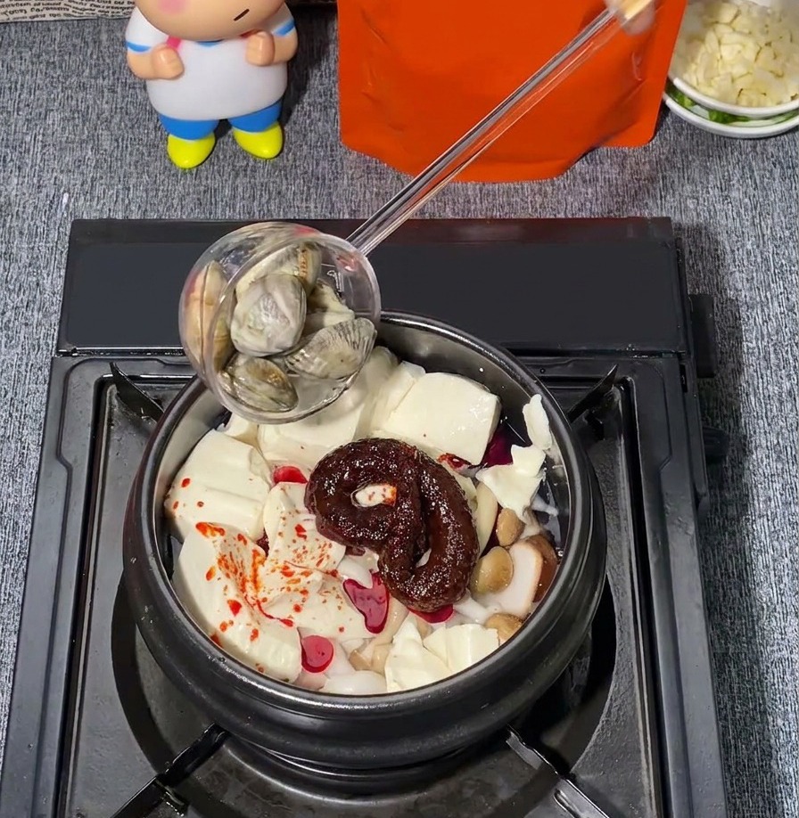 Vongole fresche aggiunte con un mestolo in una pentola di pietra contenente tofu tenero, funghi e condimento al peperoncino rosso.