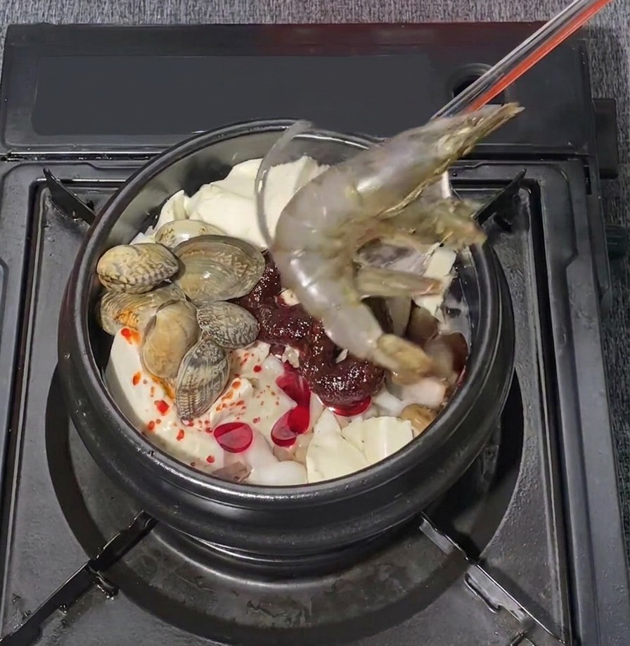 Due grandi gamberoni crudi vengono adagiati con cura in una pentola bollente di zuppa di tofu coreana ai frutti di mare.
