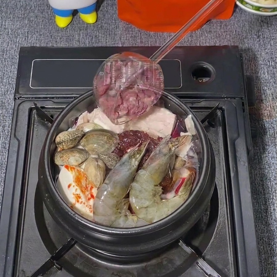 Sottili fettine di manzo crudo aggiunte da un mestolo su una pentola di stufato coreano che contiene già gamberi e vongole.