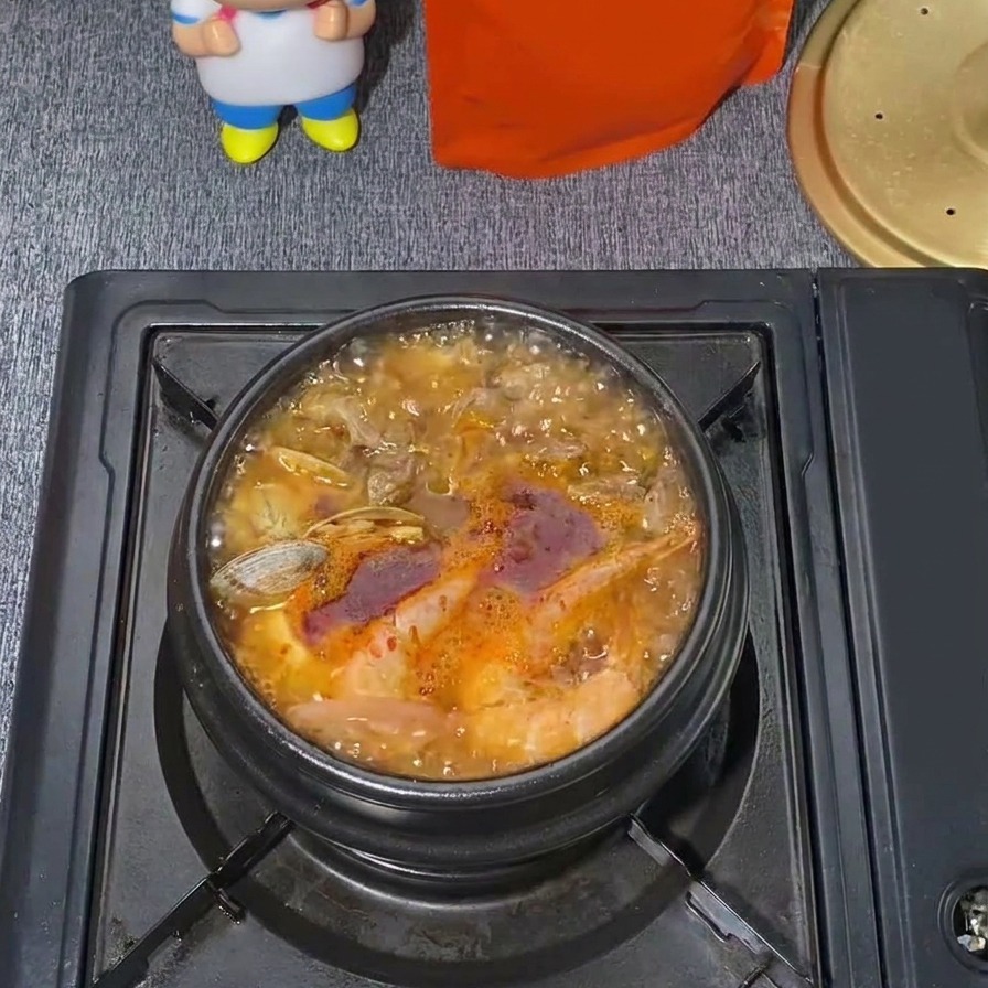 Una pentola bollente di zuppa coreana di tofu tenero ai frutti di mare con un brodo rosso vibrante, vongole visibili e vapore che sale.