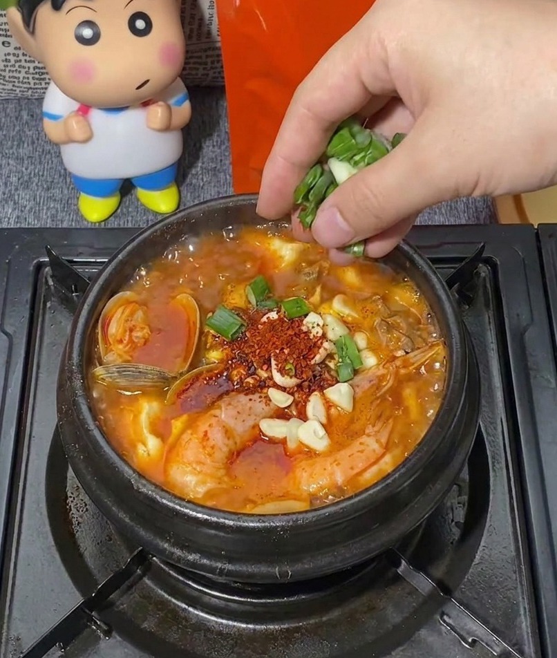 Una mano distribuisce una manciata di cipollotti verdi tritati in una pentola di pietra nera bollente di zuppa coreana con gamberi, vongole e aglio tritato.