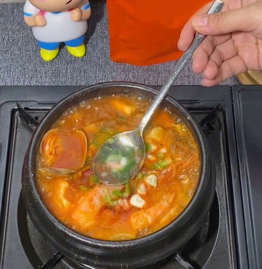 Un cucchiaio di metallo mescola i cipollotti tritati in una pentola bollente di zuppa rossa di tofu e frutti di mare.
