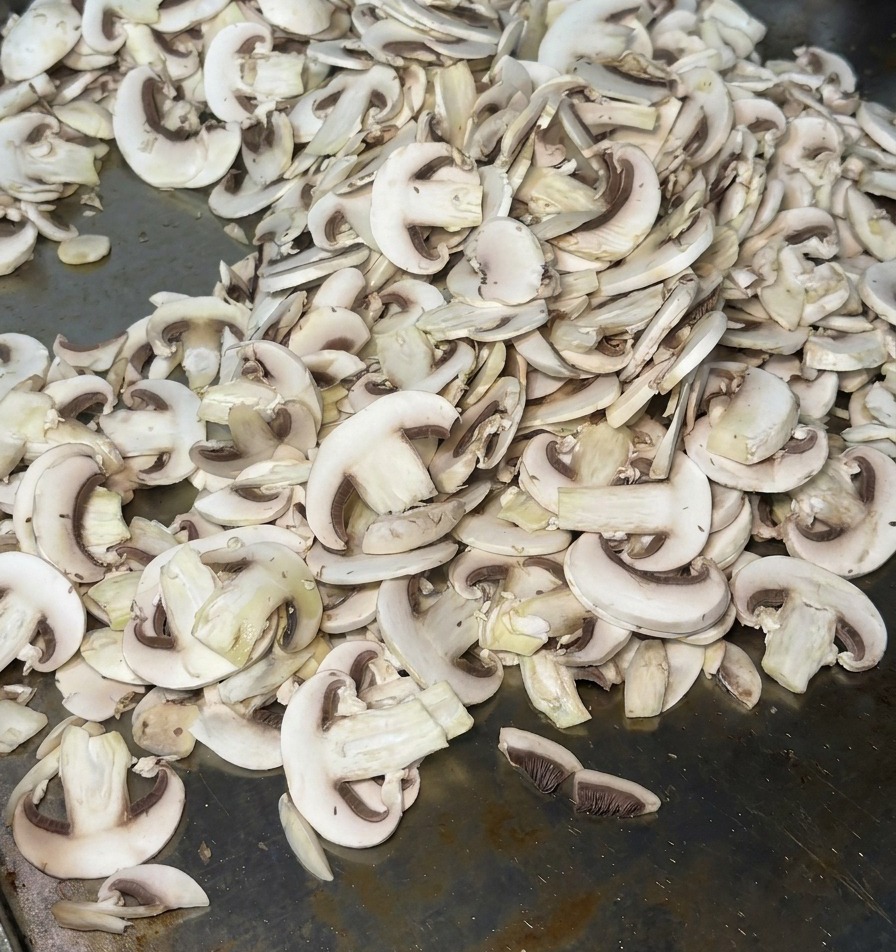 Funghi affettati finemente distribuiti su una piastra di metallo calda che rilasciano vapore allinizio della cottura.
