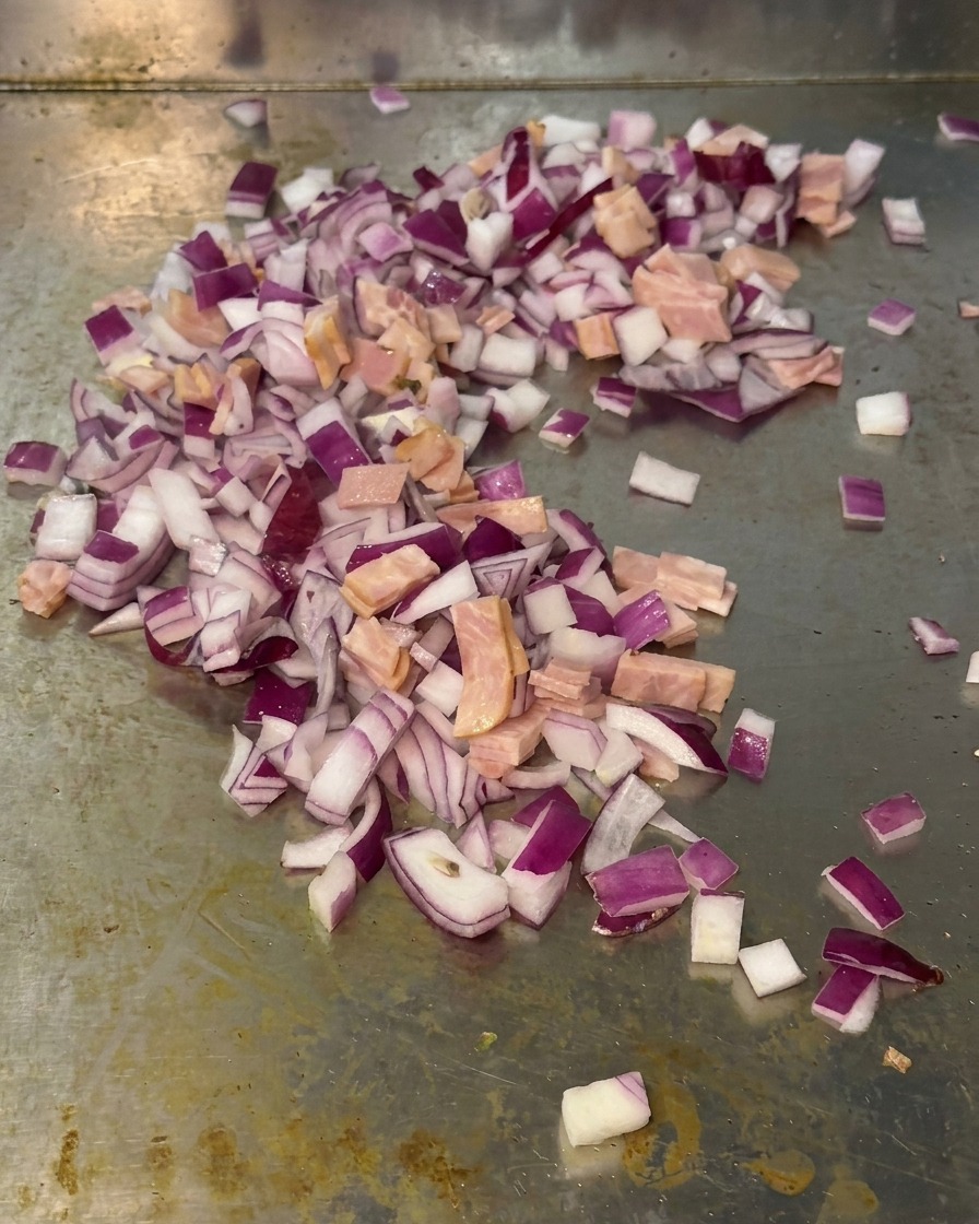 Un mix di cipolle rosse a dadini e piccoli pezzi di bacon saltati insieme sulla superficie di una piastra metallica.