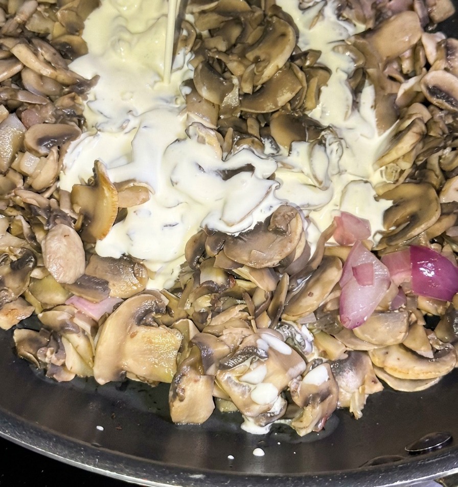 Un filo di panna fresca liquida versato da un cartone blu in una padella piena di funghi saltati e cipolle rosse a dadini.