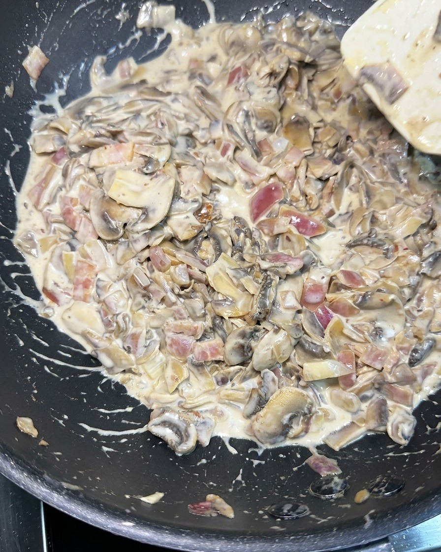 Una spatola bianca che mescola una salsa densa e cremosa ricca di funghi a fette, cipolla rossa a dadini e pezzi di bacon in una padella nera.
