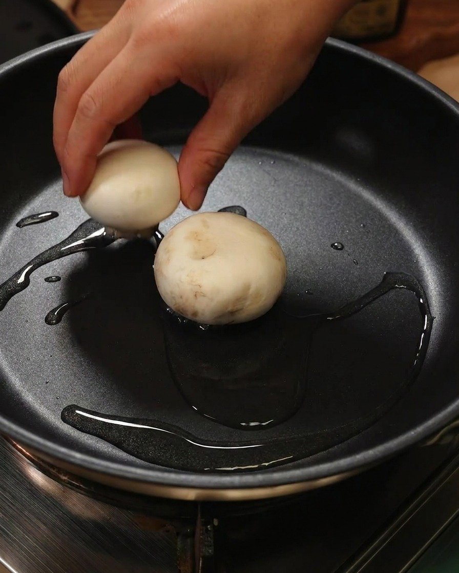 Una mano posiziona una cappella di fungo farcita con pasta di gamberi rivolta verso il basso in una padella oliata