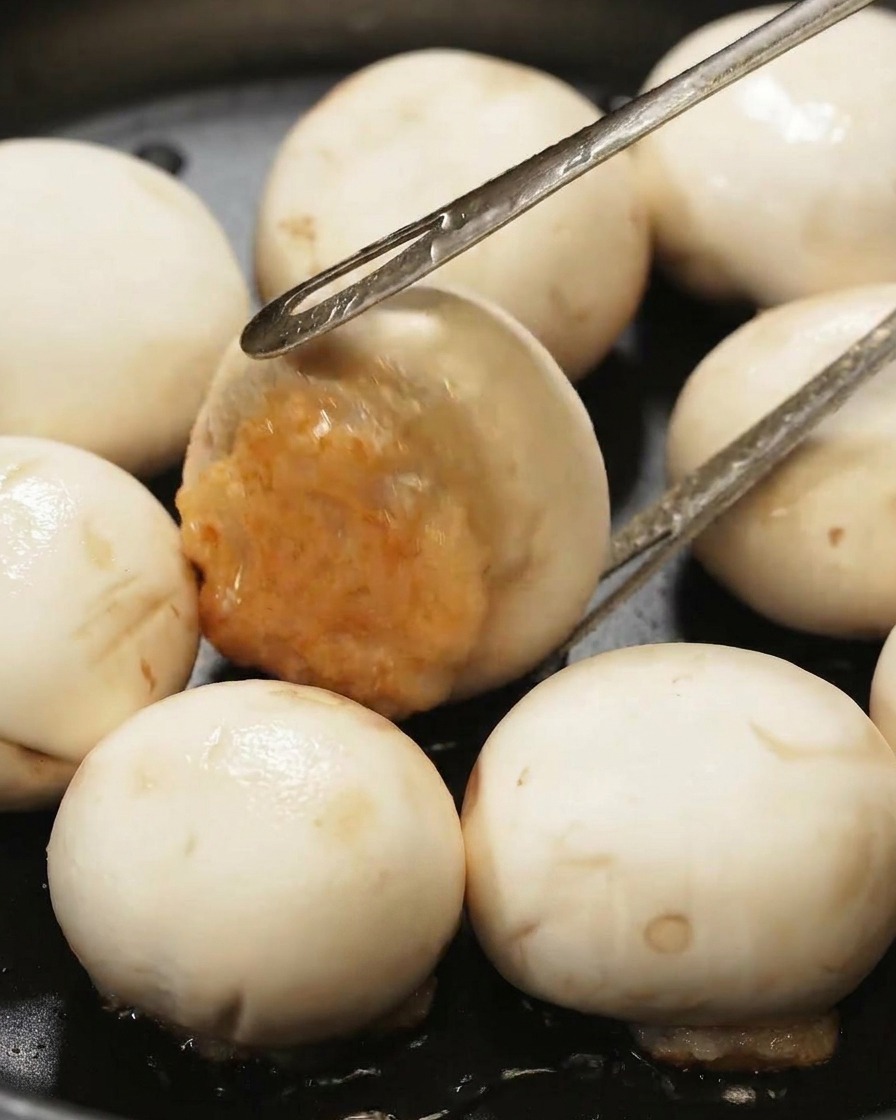 Pinze metalliche girano una cappella di fungo ripiena in una padella nera, mostrando il ripieno di gamberi ben dorato