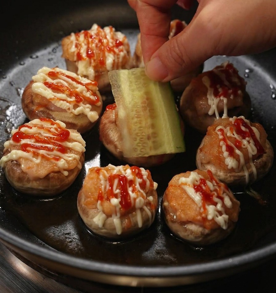 Una mano posiziona una fetta rettangolare di cetriolo fresco su un burger di funghi ripieno guarnito con maionese e ketchup
