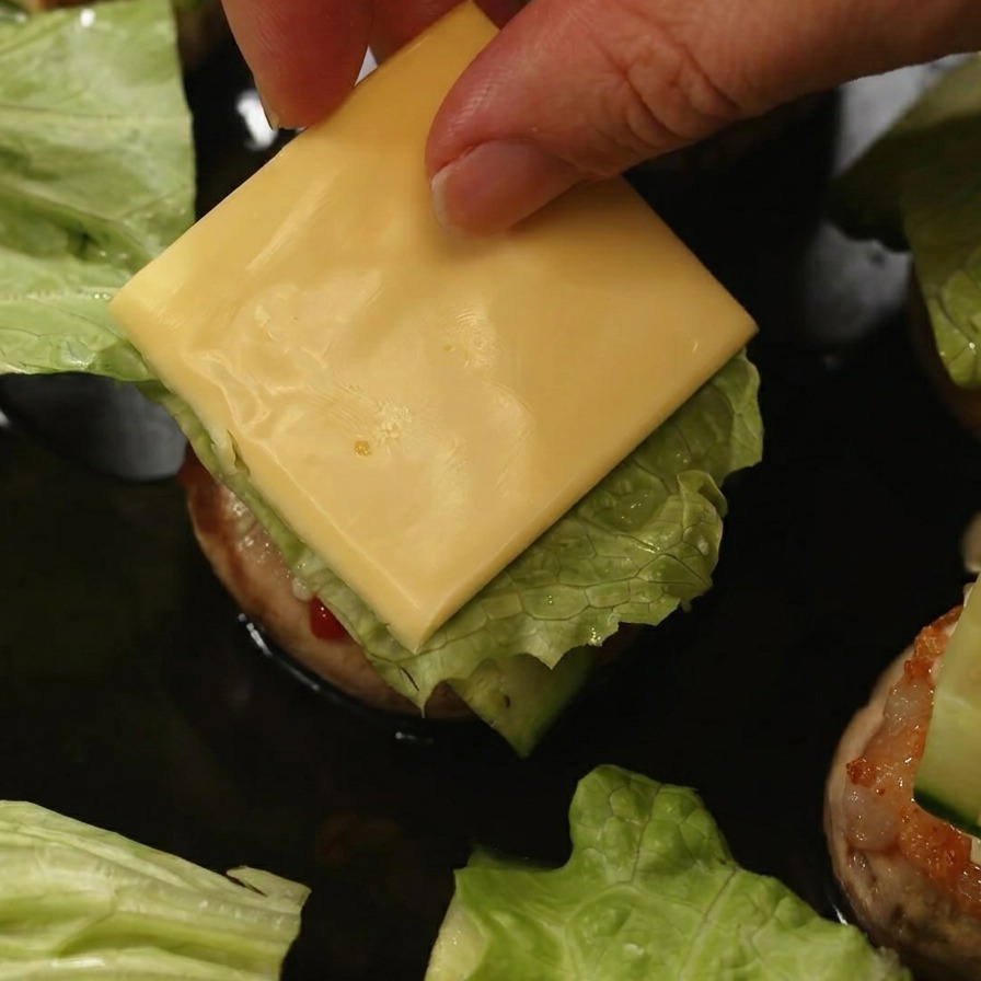Una mano posiziona una fetta quadrata di formaggio giallo sopra la lattuga e il cetriolo allinterno di un burger di funghi