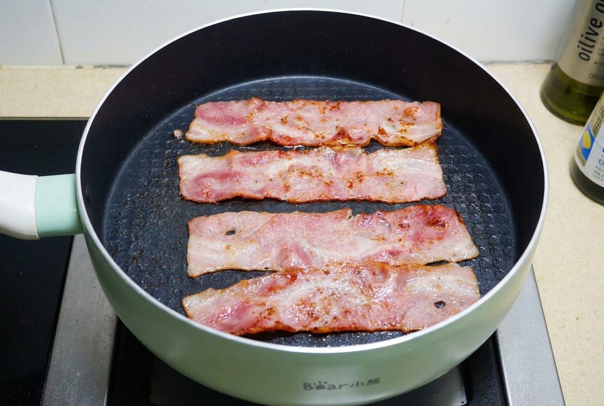 Strisce di bacon che sfrigolano in una padella antiaderente fino a diventare dorate e croccanti.