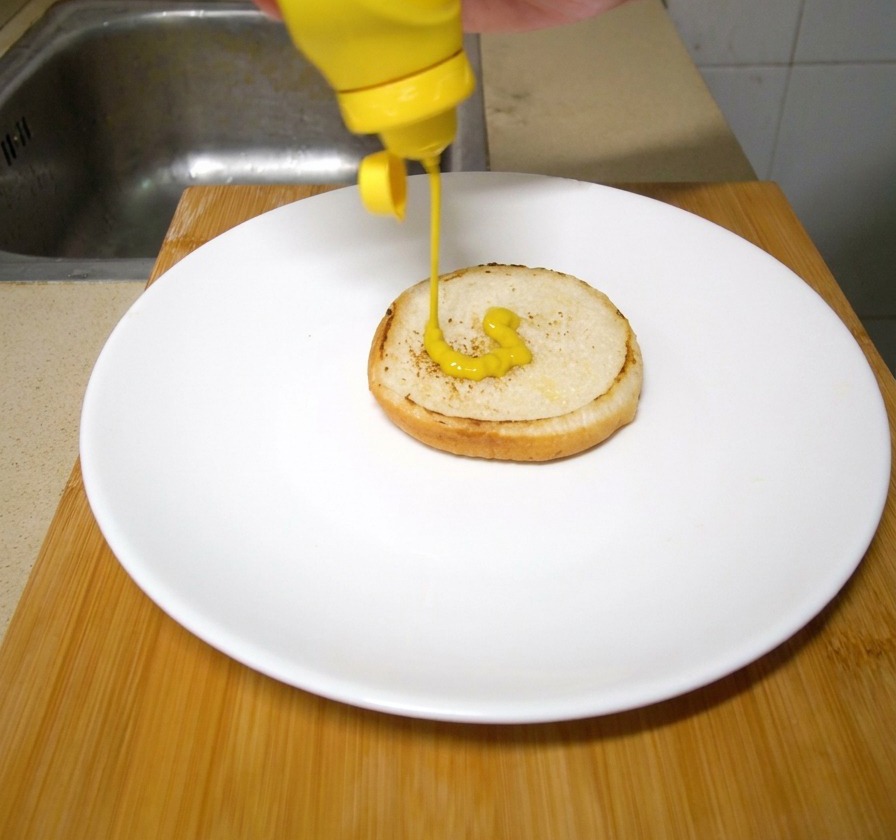 Una mano che versa della senape gialla sulla base tostata del panino per hamburger disposto su un piatto.