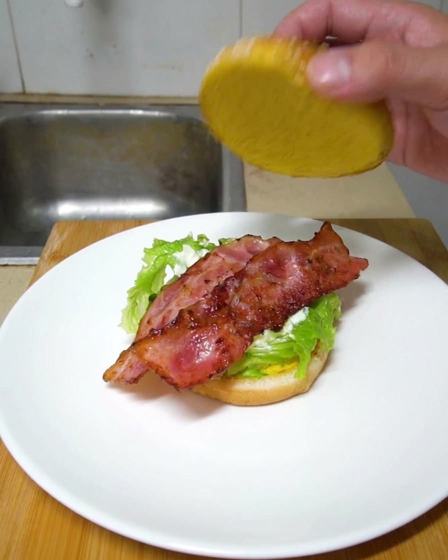 Una mano che chiude lhamburger appoggiando la parte superiore del panino sul bacon croccante.