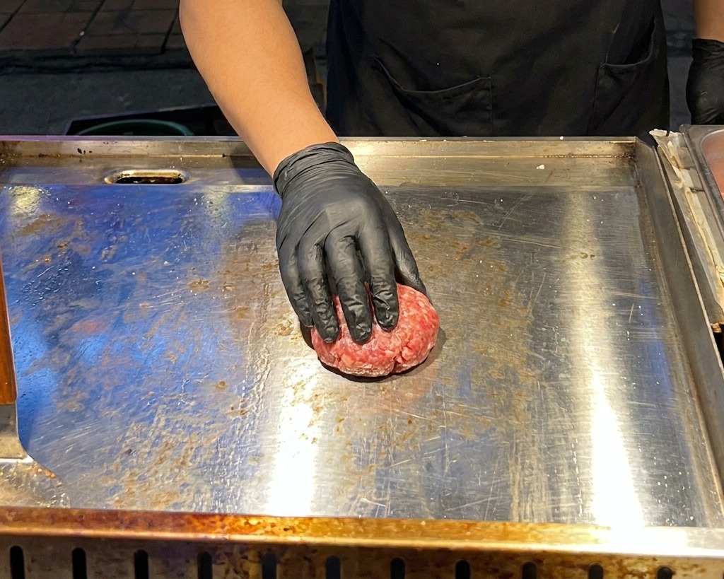 Una mano con un guanto nero usa una pressa metallica piatta per schiacciare una polpetta di manzo cruda su una piastra calda.