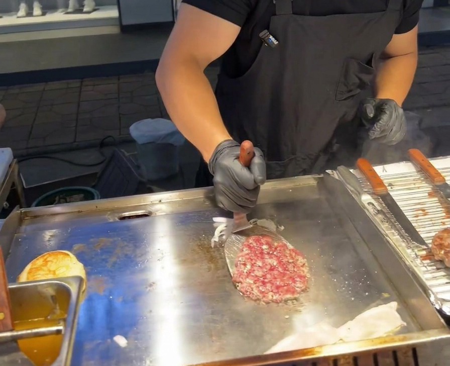 Una spatola metallica gira un patty di manzo posizionandolo sopra un mucchietto di cipolle rosse grigliate sulla piastra.