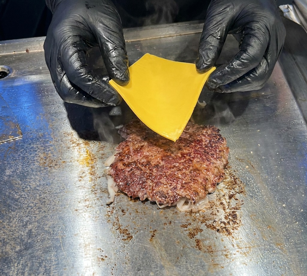 Primo piano di mani con guanti neri che adagiano con cura una fetta di cheddar giallo su un patty di manzo rosolato e sfrigolante sulla piastra.