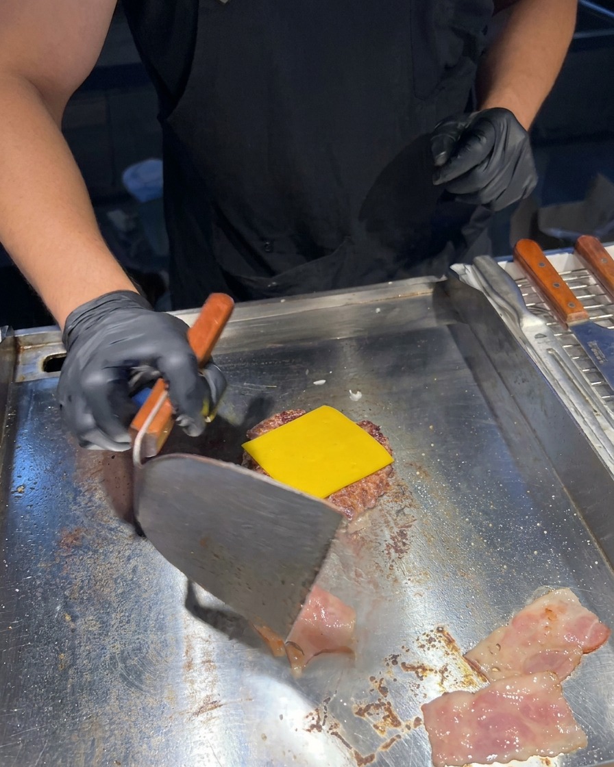 Una spatola metallica gira delle strisce sfrigolanti di bacon sulla piastra accanto al patty con il formaggio fuso.