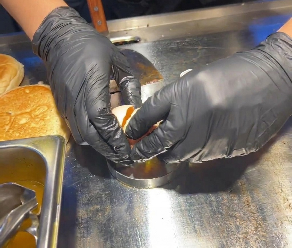 Mani con guanti neri rompono un uovo fresco allinterno di un coppapasta metallico circolare posizionato sulla piastra calda.