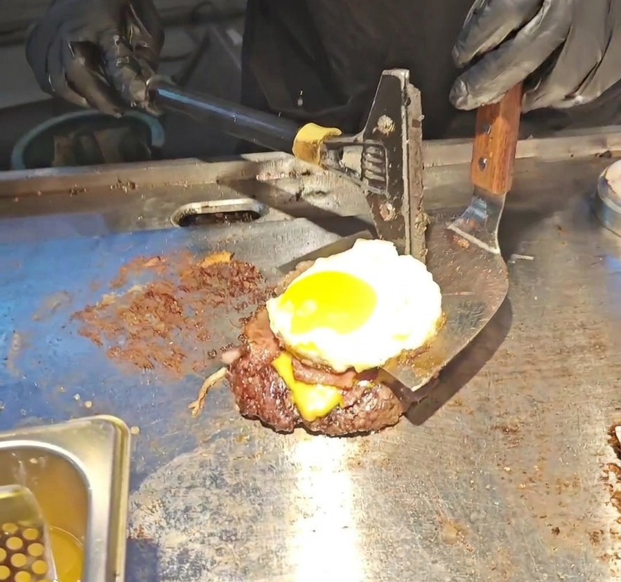 Una larga spatola metallica solleva unalta pila da burger composta da patty di manzo, formaggio fuso, bacon e un uovo allocchio di bue.
